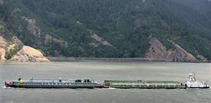 A tug pushes a wheat barge downstream providing competition with the rail line seen along the shore in the distance.